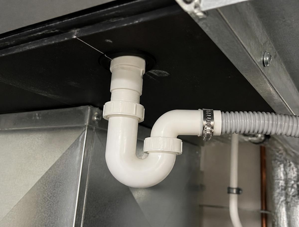 White plastic P-trap connected to the drain pan outlet beneath a ducted air handling unit and running into a flexible condensate hose