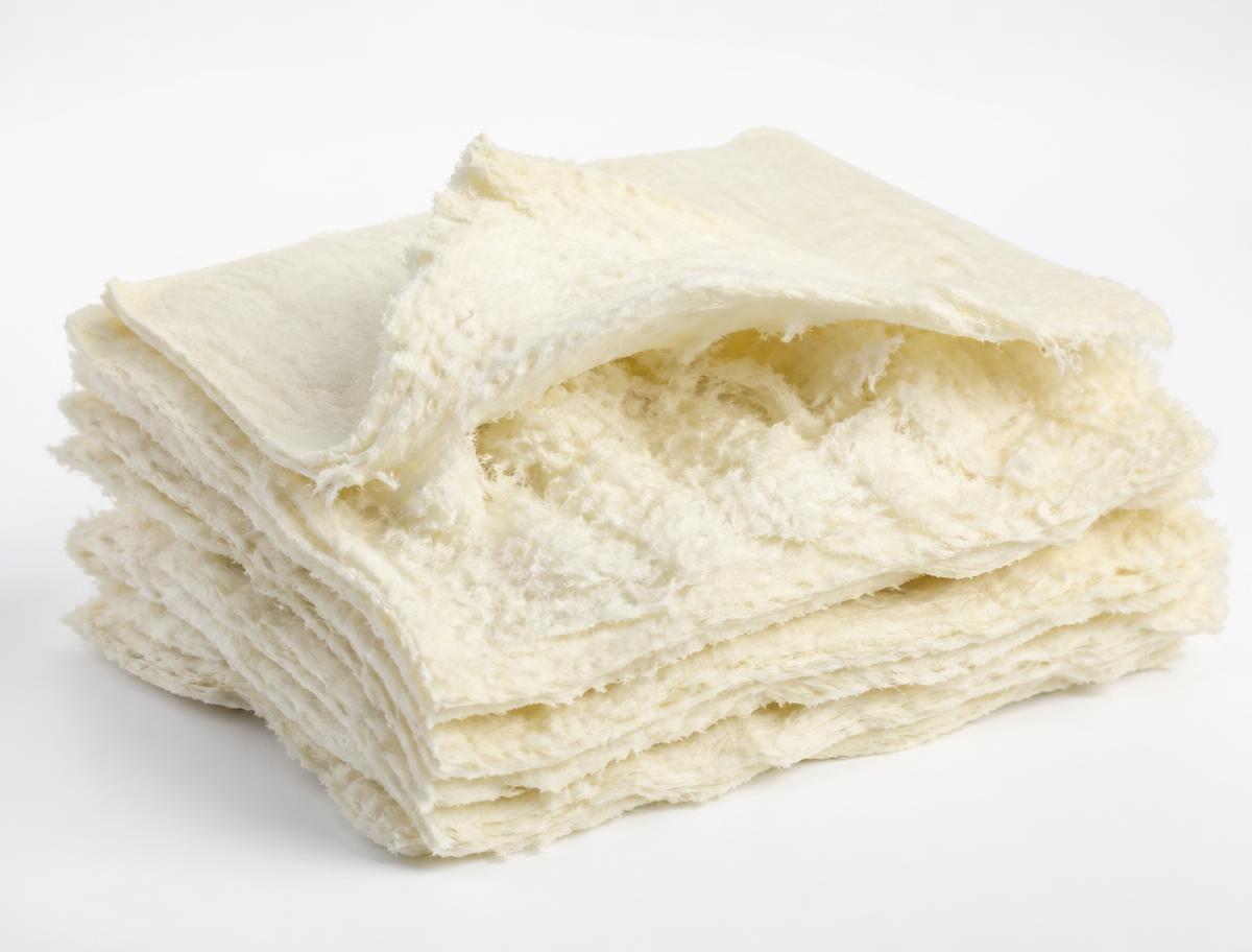 Stack of pale cream fluff pulp sheets with one sheet teased open to show fluffed absorbent cellulose fibres