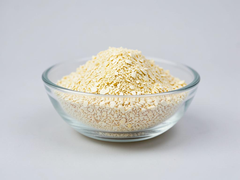 Nutritional yeast shown as pale yellow flakes heaped in a shallow glass dish in a neutral studio photograph