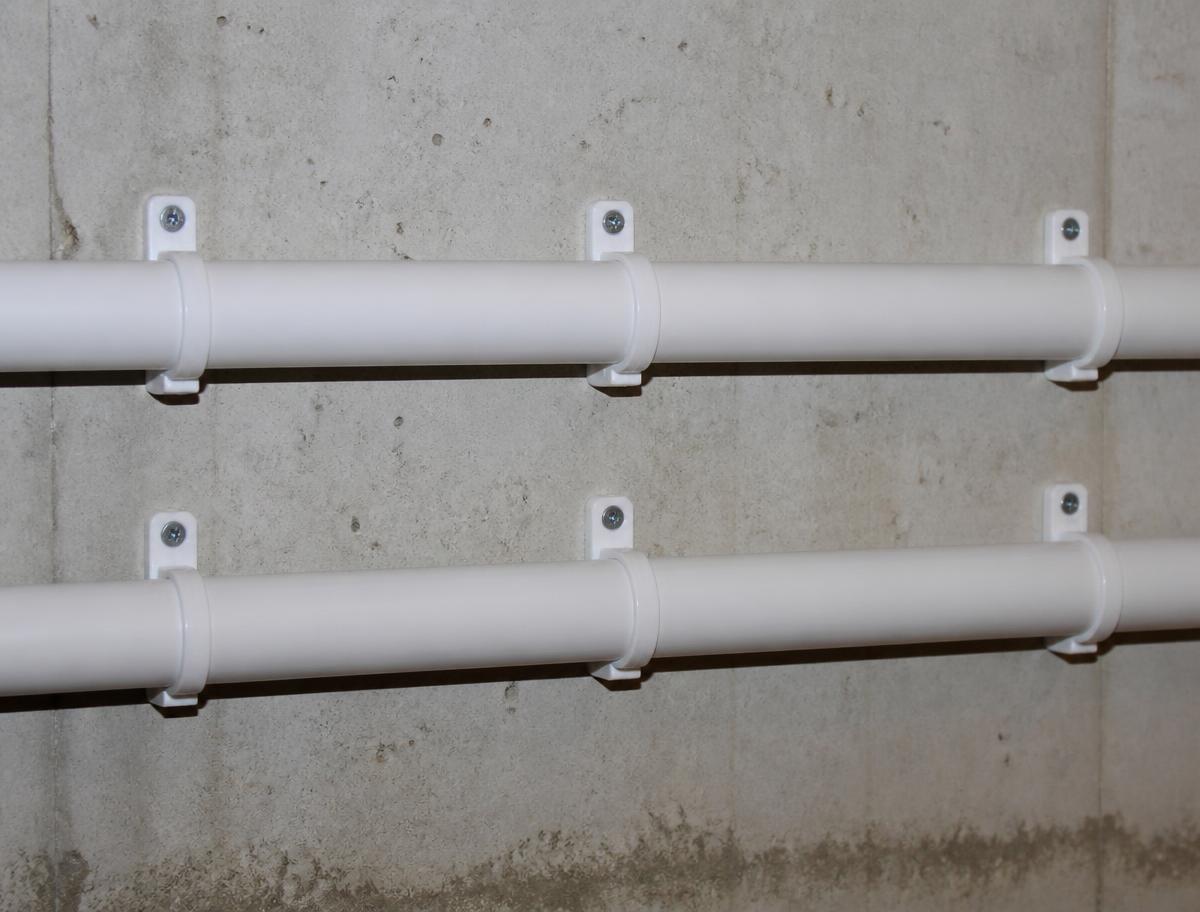 Two parallel white pipes running along a concrete wall, supported at regular spacing by screw-fixed white plastic pipe clips