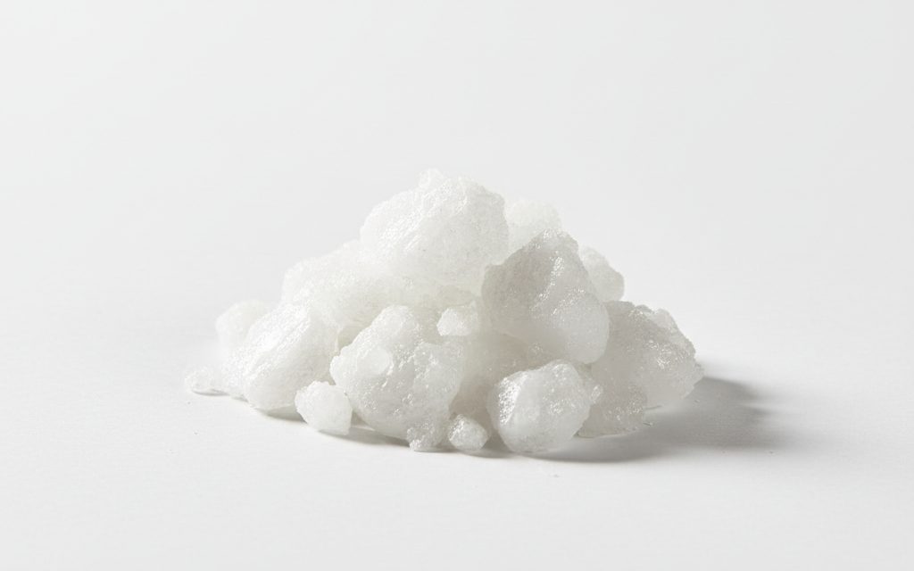 Grade confirmed at quotation — white, deliquescent solid pellets or flakes that are highly caustic and rapidly absorb moisture from the surrounding air, studio product photograph
