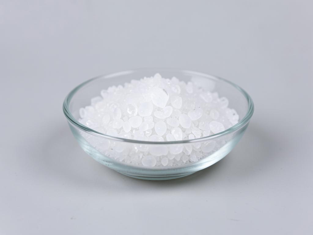 Sodium acetate shown as translucent white crystalline granules in a clear glass dish in a neutral studio photograph